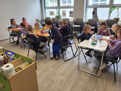 Na zdjęciu znajdują się uczestnicy zajęć. Dzieci siedzą przy stole i czytają książki. Zdjęcie wykonano w sali wielofunkcyjnej biblioteki.