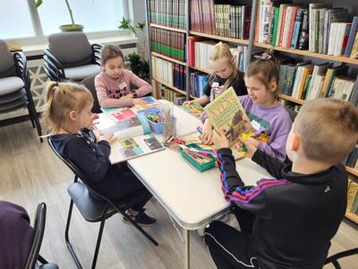 Na zdjęciu znajdują się uczestnicy zajęć. Dzieci siedzą przy stole i czytają książki. Zdjęcie wykonano w sali wielofunkcyjnej biblioteki.