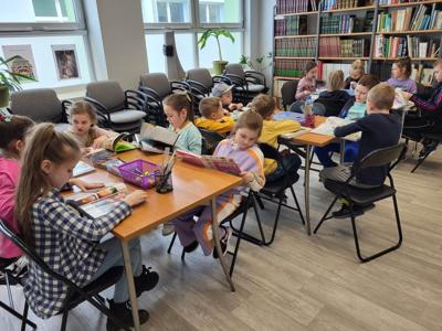 Na zdjęciu znajdują się uczestnicy zajęć. Dzieci siedzą przy stole i czytają książki. Zdjęcie wykonano w sali wielofunkcyjnej biblioteki.