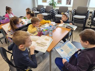 Na zdjęciu znajdują się uczestnicy zajęć. Dzieci siedzą przy stole i czytają książki. Zdjęcie wykonano w sali wielofunkcyjnej biblioteki.