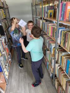 Na zdjęciu znajdują się uczestnicy warsztatów. Dzieci wykonują zadania.  W tle księgozbiór biblioteki.