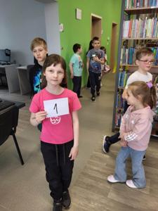 Na zdjęciu znajdują się uczestnicy warsztatów. Dzieci wykonują zadania.  W tle księgozbiór biblioteki.