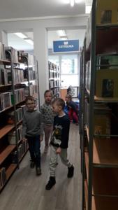 Na zdjęciu znajdują się uczestnicy zajęć. Dzieci idą pomiędzy regałami z książkami. Zdjęcie wykonano w bibliotece.