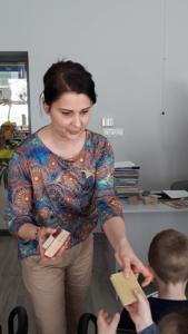 Na zdjęciu znajdują się uczestnicy zajęć. Dzieci siedzą na krzesłach. Panie stoją. Zdjęcie wykonano w sali wielofunkcyjnej biblioteki.
