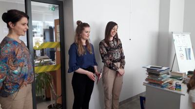 Na zdjęciu znajdują się uczestnicy zajęć. Dzieci siedzą na krzesłach. Panie stoją. Zdjęcie wykonano w sali wielofunkcyjnej biblioteki.