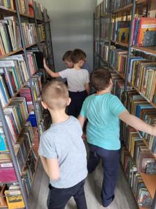 Na zdjęciu znajdują się uczestnicy zajęć. Dzieci idą pomiędzy regałami z książkami. Zdjęcie wykonano w bibliotece.