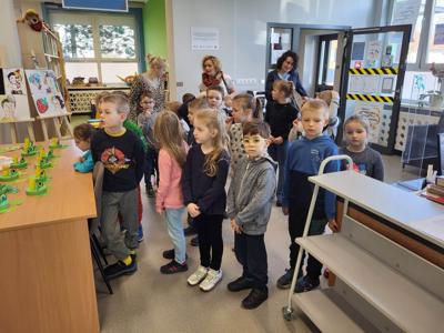 Na zdjęciu znajdują się uczestnicy zajęć. Dzieci stoją przy stole. Zdjęcie wykonano w bibliotece.