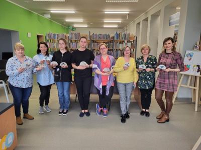 Na zdjęciu znajdują się uczestnicy warsztatów. Uczestniczki trzymają w rękach serwetniki. Zdjęcie wykonano w bibliotece.