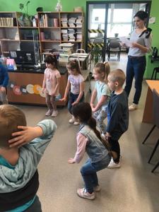 Na zdjęciu znajdują się uczestnicy zajęć. Dzieci stoją z kółku i wykonują zadania. Zdjęcie wykonano w bibliotece.