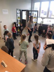 Na zdjęciu znajdują się uczestnicy zajęć. Dzieci stoją z kółku i wykonują zadania. Zdjęcie wykonano w bibliotece.