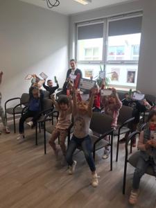 Na zdjęciu znajdują się uczestnicy zajęć. Dzieci siedzą na krzesłach. Zdjęcie wykonano w bibliotece.