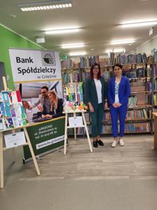 Na zdjęciu znajdują się dwie osoby. W tle księgozbiór biblioteki, prace dzieci i baner Banku Spółdzielczego.