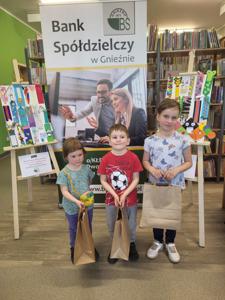Na zdjęciu znajdują się uczestnicy konkursu. W tle księgozbiór biblioteki, prace dzieci i baner Banku Spółdzielczego.