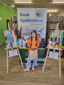 Na zdjęciu znajdują się uczestnicy konkursu. W tle księgozbiór biblioteki, prace dzieci i baner Banku Spółdzielczego.