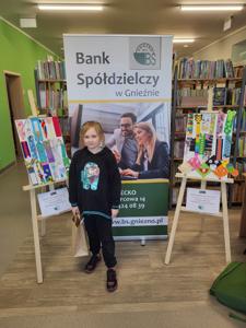 Na zdjęciu znajdują się uczestnicy konkursu. W tle księgozbiór biblioteki, prace dzieci i baner Banku Spółdzielczego.