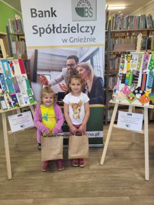Na zdjęciu znajdują się uczestnicy konkursu. W tle księgozbiór biblioteki, prace dzieci i baner Banku Spółdzielczego.