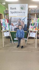 Na zdjęciu znajdują się uczestnicy konkursu. W tle księgozbiór biblioteki, prace dzieci i baner Banku Spółdzielczego.