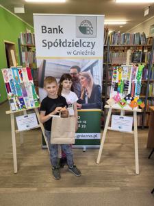 Na zdjęciu znajdują się uczestnicy konkursu. W tle księgozbiór biblioteki, prace dzieci i baner Banku Spółdzielczego.