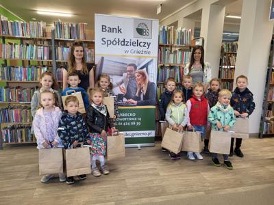 Na zdjęciu znajdują się uczestnicy konkursu. W tle księgozbiór biblioteki, prace dzieci i baner Banku Spółdzielczego.