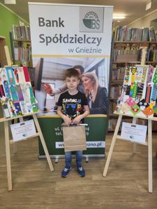 Na zdjęciu znajdują się uczestnicy konkursu. W tle księgozbiór biblioteki, prace dzieci i baner Banku Spółdzielczego.