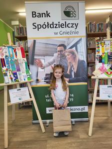 Na zdjęciu znajdują się uczestnicy konkursu. W tle księgozbiór biblioteki, prace dzieci i baner Banku Spółdzielczego.