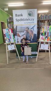 Na zdjęciu znajdują się uczestnicy konkursu. W tle księgozbiór biblioteki, prace dzieci i baner Banku Spółdzielczego.