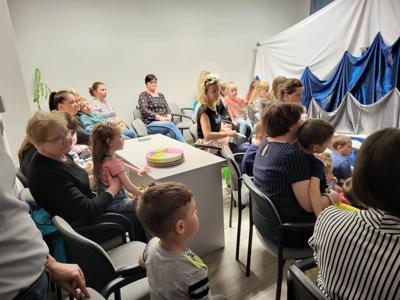 Na zdjęciu znajdują się uczestnicy przedstawienia. Dzieci i rodzice siedzą na krzesłach i poduszkach. W tle scenka teatralna.