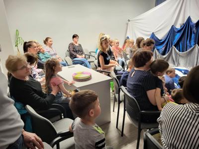 Na zdjęciu znajdują się uczestnicy przedstawienia. Dzieci i rodzice siedzą na krzesłach i poduszkach. W tle scenka teatralna.