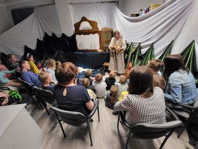 Na zdjęciu znajdują się uczestnicy przedstawienia. Dzieci i rodzice siedzą na krzesłach i poduszkach. W tle scenka teatralna.