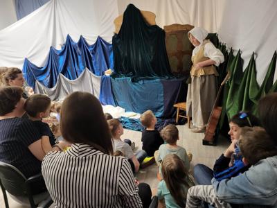 Na zdjęciu znajdują się uczestnicy przedstawienia. Dzieci i rodzice siedzą na krzesłach i poduszkach. W tle scenka teatralna.