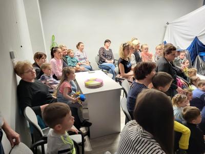 Na zdjęciu znajdują się uczestnicy przedstawienia. Dzieci i rodzice siedzą na krzesłach i poduszkach. W tle scenka teatralna.