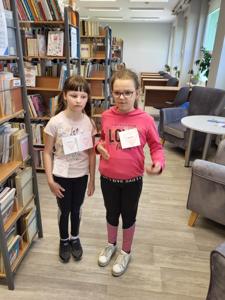 Na zdjęciu znajdują się uczestnicy zajęć. Dzieci stoją przy regałach. Zdjęcie wykonano w bibliotece.