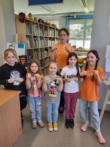 Na zdjęciu znajdują się uczestnicy warsztatów biżuteryjnych. Młodzież trzyma w ręce kolczyki. W tle księgozbiór biblioteki.