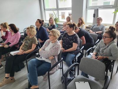 Na zdjęciu znajdują się uczestnicy warsztatów. Uczestnicy siedzą na krzesłach. Prowadząca wyświetla prezentację multimedialną.