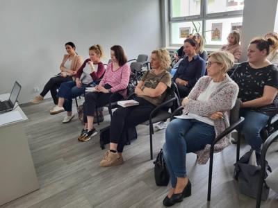 Na zdjęciu znajdują się uczestnicy warsztatów. Uczestnicy siedzą na krzesłach. Prowadząca wyświetla prezentację multimedialną.