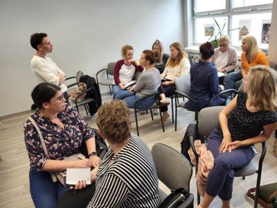 Na zdjęciu znajdują się uczestnicy warsztatów. Uczestnicy siedzą na krzesłach. Prowadząca wyświetla prezentację multimedialną.