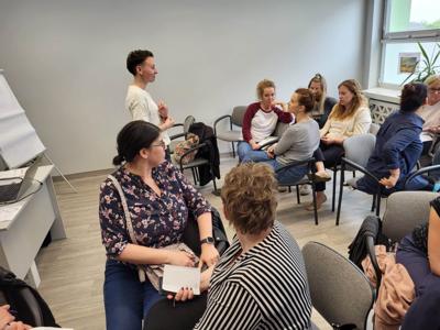 Na zdjęciu znajdują się uczestnicy warsztatów. Uczestnicy siedzą na krzesłach. Prowadząca wyświetla prezentację multimedialną.