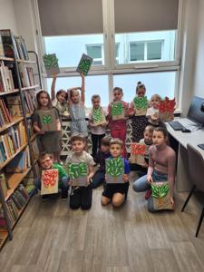 Na zdjęciu znajdują się uczestnicy warsztatów. Dzieci trzymają w rękach prace plastyczne. Zdjęcie wykonano w bibliotece.