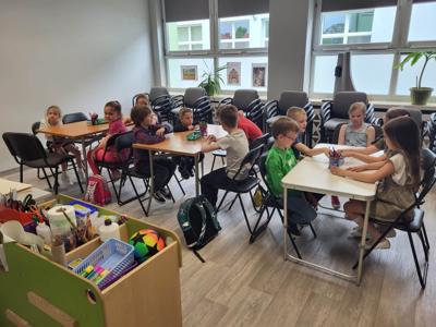 Na zdjęciu znajdują się uczestnicy warsztatów. Dzieci siedzą przy stole na krzesłach. Zdjęcie wykonano w bibliotece.