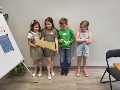 Na zdjęciu znajdują się uczestnicy warsztatów. Dzieci stoją i trzymają rękach kartkę. Zdjęcie wykonano w bibliotece.