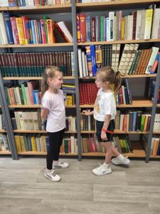 Na zdjęciu znajdują się uczestnicy warsztatów. Dzieci stoją i wykonują zadania. W tle księgozbiór biblioteki. Zdjęcie wykonano w bibliotece.