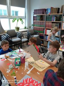 Na zdjęciu znajdują się uczestnicy warsztatów. Dzieci siedzą na krzesłach i wykonują prace plastyczne. Zdjęcie wykonano w bibliotece.