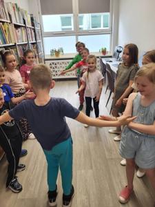 Na zdjęciu znajdują się uczestnicy warsztatów. Dzieci stoją i wykonują zadania. Zdjęcie wykonano w bibliotece.