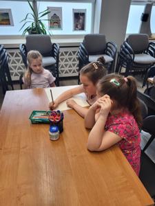 Na zdjęciu znajdują się uczestnicy warsztatów. Dzieci siedzą na krzesłach i wykonują prace plastyczne. Zdjęcie wykonano w bibliotece.