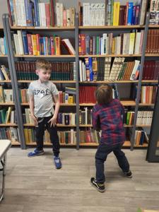Na zdjęciu znajdują się uczestnicy warsztatów. Dzieci stoją i wykonują zadania. W tle księgozbiór biblioteki. Zdjęcie wykonano w bibliotece.