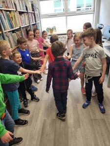 Na zdjęciu znajdują się uczestnicy warsztatów. Dzieci stoją i wykonują zadania. Zdjęcie wykonano w bibliotece.