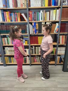 Na zdjęciu znajdują się uczestnicy warsztatów. Dzieci stoją i wykonują zadania. W tle księgozbiór biblioteki. Zdjęcie wykonano w bibliotece.
