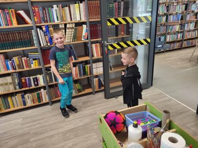 Na zdjęciu znajdują się uczestnicy warsztatów. Dzieci stoją i wykonują zadania. W tle księgozbiór biblioteki. Zdjęcie wykonano w bibliotece.
