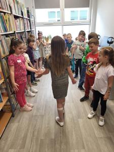 Na zdjęciu znajdują się uczestnicy warsztatów. Dzieci stoją i wykonują zadania. Zdjęcie wykonano w bibliotece.