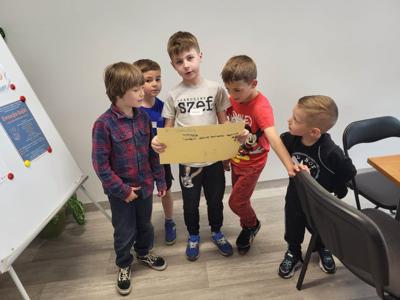Na zdjęciu znajdują się uczestnicy warsztatów. Dzieci stoją i trzymają rękach kartkę. Zdjęcie wykonano w bibliotece.